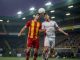 Two opposing footballers—one in red-yellow, one in white with red accents—challenge for a high